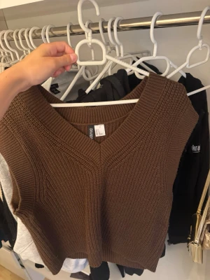 Brun stickad väst med v-ringning - Säljer en brun stickad väst med bred v-ringning och ribbade detaljer. Västen är ärmlös och har en rak, något kortare passform. Perfekt att styla över en skjorta eller t-shirt för en trendig lager-på-lager-look.