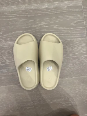 Beige slides från Adidas - Snygga beigea tofflor från Adidas med minimalistisk design och bred rem över foten. Tillverkade i ett mjukt och lätt material med räfflad sula för extra grepp. Perfekta för chill dagar hemma eller till och från gymmet.