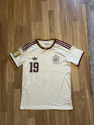 Spansk landslagströja Adidas nr 19 - Säljer en vit Spanien fotbollströja från Adidas med vinröda detaljer och nummer 19. Tröjan har Lamine Yamal tryckt på ryggen, klassiska Adidas-ränder på axlarna och landslagets emblem på bröstet. Tillverkad i lätt och ventilerande material, perfekt för match eller träning.