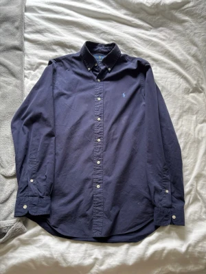 Mörkblå skjorta från Ralph Lauren - Klassisk mörkblå skjorta från Ralph Lauren med button-down krage och broderad logga på bröstet. Skjortan har långa ärmar, vita knappar och är tillverkad i mjuk bomull