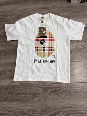 Vit BAPE t-shirt med tryck - Säljer en vit t-shirt från BAPE med det klassiska apansiktet i ett rutigt mönster på bröstet och texten 'BY BATHING APE' under. T-shirten har rund hals och korta ärmar. Perfekt för dig som gillar streetwear och vill sticka ut.