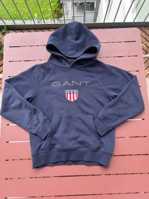 Blå hoodie från GANT med logga - Mörkblå hoodie från GANT med broderad logga och amerikanskinspirerat sköldmärke på bröstet. Klassisk känguruficka framtill och huva. Perfekt för en avslappnad och stilren look. Tillverkad i mjukt bomullsmaterial.