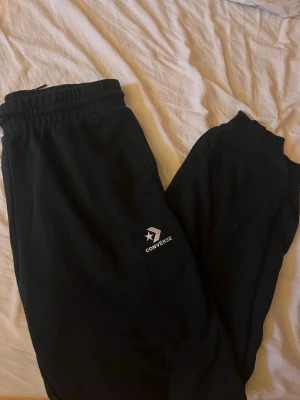 Svarta mjukisbyxor från Converse - Säljer ett par svarta mjukisbyxor från Converse med logga på benet. Byxorna har resår i midjan och vid bensluten för en skön och avslappnad passform. Perfekta för chill dagar eller när du vill vara bekväm.