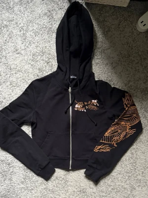 Svart hoodie med draktryck och dragkedja - Cool svart hoodie från Bershka med orange drakmotiv på ärm och rygg samt texten 'Energy Flow'. Croppad modell med huva, dragkedja och fickor framtill. Perfekt för dig som gillar streetstyle och unika detaljer.