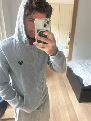Comme des carcobs hoodie - Snygg grå hoodie med dragkedja och huva. På bröstet och ryggen finns en svart hjärtdetalj med ögon, vilket ger en unik och trendig look. Hoodien har fickor framtill och är perfekt för en avslappnad stil.