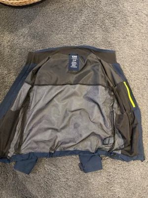 Blå vindjacka från Helly Hansen - Snygg blå vindjacka från Helly Hansen med meshfoder och detaljer i gult. Jackan har regular fit och är tillverkad i slitstarkt syntetmaterial. Perfekt för blåsiga dagar och outdoor-aktiviteter. Praktiska fickor och justerbara ärmslut. Super skön och bra mot regn men också väldigt snygg. Små tecken på använding lite här och var. Är 175 och 65kg o sitter bra på mig.