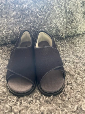 Svarta sandaler med kardborreband - Säljer ett par svarta sandaler med breda kardborreband och öppen tå. De har en mjuk ovandel i textil och en stadig sula i gummi. Perfekta för dig som vill ha bekväma och lättanvända skor med enkel design.