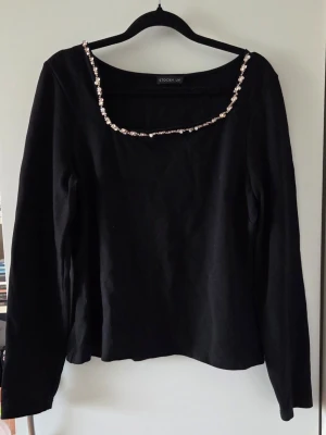 Svart topp med pärldetaljer - Snygg svart långärmad topp med pärlor längs den assymetriska ringningen. Enkel men elegant design som passar till många olika looks. Tillverkad i mjukt material och har en normal passform. Det saknas tyvärr några pärlor, men inget man tänker på direkt.
