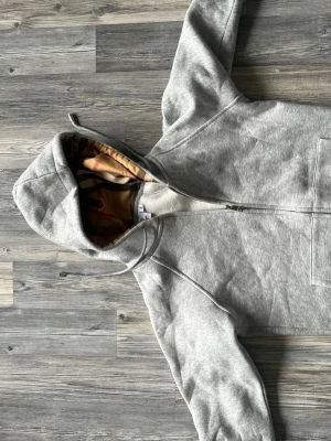Grå zip hoodie burberry  - Säljer en grå hoodie med dragkedja och huva. Insidan av huvan har ett beige och rödbrunt mönstrat foder som ger en unik touch. Hoodien har fickor framtill och är gjord i mjukt sweatshirtmaterial. Perfekt för chill dagar eller när du vill ha en clean look. Jätteliten i storlek 