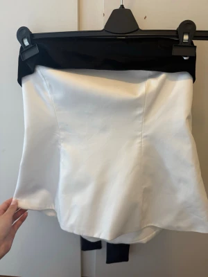 Vit axelbandslös topp från Zara med svart rosett - Säljer en elegant vit axelbandslös topp från Zara med en bred svart rosett i midjan. Toppen har en bandeauformad överdel och är perfekt för dig som vill ha en stilren och trendig look. Materialet känns slätt och har en lätt glans.
