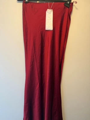 Röd långkjol i satin från Gina Tricot - Snygg vinröd långkjol i satin med enkel och stilren design. Kjolen har en lätt glansig yta och en rak passform som ger en elegant look. Perfekt för dig som vill sticka ut med en färgstark och trendig kjol.
