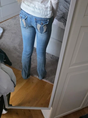 Miss me jeans - Säljer ett par blå bootcut jeans från Miss Me med slitningar framtill och unika diamant fickor bak. Jeansen har låg midja och snygga kontrastsömmar. Perfekta för dig som vill ha ett par jeans med coola detaljer och avslappnad vibe.
