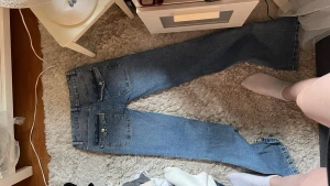 Blå raka jeans med dragkedjor - Säljer ett par blå jeans med raka ben och diamantliknande stenar på bakfickorna. Jeansen har klassisk femficksdesign och är tillverkade i denim. Perfekta för en avslappnad och trendig look. Står 40 i storlek men jag har sytt upp dom till mer lågmidjade så storleken har blivit mindre med de.