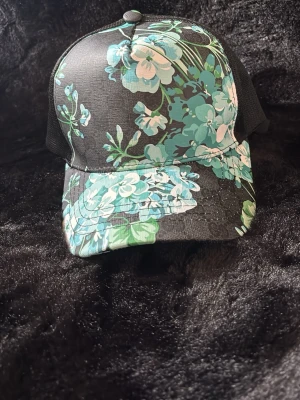 Gucci blommig keps svart/grön - Snygg keps från Gucci med svart bas och blommigt mönster i grönt, turkost och vitt. Kepsen har klassisk form med böjd skärm och mesh på sidorna. Insidan är märkt med Gucci-logga. Perfekt för dig som vill sticka ut med en unik accessoar.