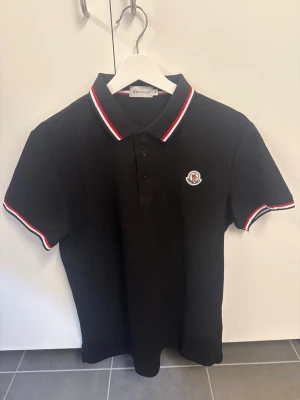 Svart Moncler pikétröja XL (sitter som L) - Klassisk svart pikétröja från Moncler med vit och röd rand på krage och ärmslut. Tröjan har knappslå framtill och Moncler-logga broderad på bröstet. Perfekt för en stilren och sportig look.