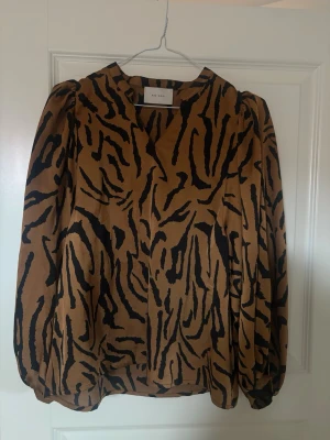 Brun zebramönstrad blus Neo Noir - Snygg brun blus från Neo Noir med svart zebramönster. Blusen har långärmade puffärmar och en liten v-ringning vid halsen. 