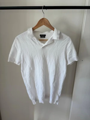 Vit stickad tenniströja - Vit stickad polotopp med klassisk krage och korta ärmar. Toppen har en enkel och stilren design som passar perfekt till jeans eller kjol. Materialet känns mjukt och luftigt, perfekt för varmare dagar.