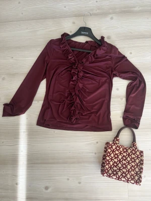 Vinröd blus med volangdetaljer - Säljer en vinröd blus med långa ärmar och snygga volangdetaljer längs ringningen och framtill. Blusen har en feminin och trendig look, perfekt att styla med jeans eller kjol. Materialet känns mjukt och följsamt mot huden.
