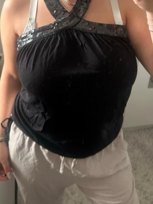 Svart topp med paljettdetalj - Snygg svart topp med breda axelband som korsas framtill och är dekorerade med glittriga paljetter. Toppen har en lös passform och är perfekt för dig som vill sticka ut lite extra. Materialet känns mjukt och bekvämt mot huden.