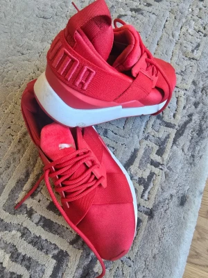 Röda sneakers från Puma - Snygga röda sneakers från Puma med vit sula och bred resårdetalj över foten. Skorna har en modern design med tydlig Puma-logga på hälen och ovansidan. Tillverkade i textil och syntetmaterial för en sportig look.