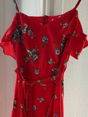 Röd blommig omlottklänning - Snygg röd omlottklänning med blå och vita blommor. Klänningen har smala axelband, volangdetaljer vid ärmarna och ett matchande knytband i midjan. Perfekt för sommarens alla tillfällen.