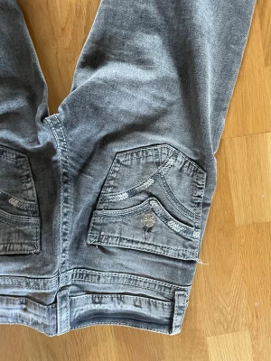 Grå bootcut jeans med fickdetaljer - Säljer ett par grå bootcut jeans med snygga detaljer på bakfickorna och slitningar för en cool look. Jeansen har normal midja och klassisk femficksmodell. Materialet är mjukt denim som ger en avslappnad känsla.(De har tappat en knapp på fickan men tycker personligen att det är lite coolt) 