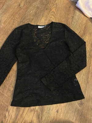 Svart spetsblus med v-ringning - Snygg svart blus i spets med lång ärm och djup v-ringning. Transparent material med blommigt mönster ger en trendig look. Perfekt att styla lager på lager eller med ett linne under.