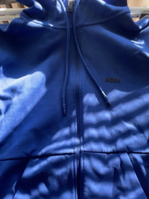 Blå hoodie med dragkedja från BOSS - Snygg blå hoodie från BOSS med dragkedja framtill och justerbar huva. Hoodien har diskret BOSS-logga på bröstet och är tillverkad i mjukt material. Perfekt för en avslappnad och stilren look. Storlek S använd ett fåtal gånger. 