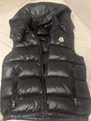 Svart dunväst från Moncler - Snygg svart dunväst från Moncler med glansig finish och hög krage. Västen har två dragkedjefickor fram, Moncler-logga på bröstet och en rejäl huva. Perfekt för lager på lager och streetwear-vibe.