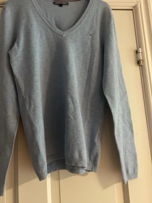 Ljusblå stickad tröja Tommy Hilfiger - Ljusblå stickad tröja från Tommy Hilfiger med V-ringning och diskret broderad logga på bröstet. Tröjan har långa ärmar och ribbade muddar vid ärmslut och nederkant. Perfekt för lager på lager och enkel att matcha.