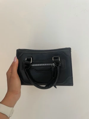 Svart liten handväska med dragkedja från SHEIN - Snygg svart handväska i kompakt modell med två handtag och en silverfärgad dragkedja framtill. Väskan har en stilren, boxig form och är tillverkad i ett glansigt material som liknar konstläder. Perfekt för dig som gillar minimalistisk och trendig design.