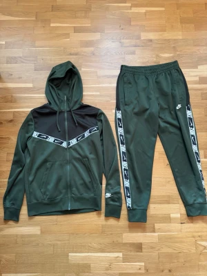 Nike grön tracksuit med huva - Snygg grön tracksuit från Nike med svart panel och vita loggor på både jacka och byxor. Jackan har dragkedja, huva och fickor, medan byxorna har resår i midjan och muddar vid benslut. Sidorna pryds av vita Nike-band med logga. Perfekt för chill eller träning.