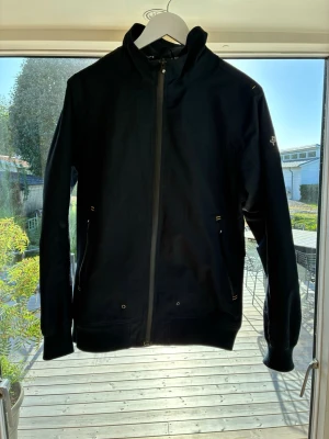 Marinblå seglarjacka från Pelle P - Snygg marinblå seglarjacka med hel dragkedja framtill. Den är vattentät och vindtät. Två fickor med dragkedja och diskreta detaljer i gul. Jackan har en gul etikett i nacken på baksidan och ribbade muddar vid ärmslut och nederkant. Perfekt för blåsiga dagar men även en lite kallare sommardag. En perfekt allround jacka helt enkelt