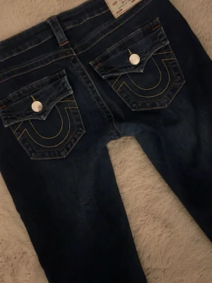 Mörkblå bootcut jeans True Religion - Säljer ett par mörkblå bootcut jeans från True Religion med kontrastsömmar och ikoniska fickdetaljer bak. Jeansen har låg midja, klassisk femficksmodell och är tillverkade i denim. W27