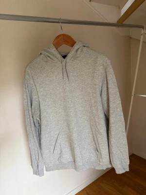 Ljusgrå hoodie med magficka - En stilren ljusgrå hoodie med huva och dragsnören. Hoodien har en klassisk magficka framtill och ribbade muddar vid ärmslut och nederkant. Tillverkad i mjukt sweatshirtmaterial som passar perfekt för en avslappnad look.
