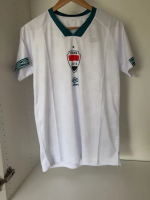 Vit Irak fotbollströja från Umbro - Säljer en vit fotbollströja från Umbro med Iraks landslagsemblem på bröstet. Tröjan har gröna detaljer vid krage och ärmslut samt diskret mönster i tyget. Perfekt för dig som vill stötta Irak på match eller träning.