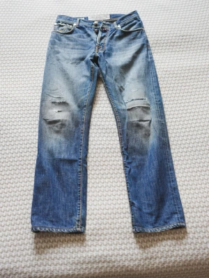 Jacob Cohën - Säljer ett par blå jeans med raka ben och slitningar över knäna. Jeansen har klassisk femficksmodell och knappgylf. Materialet är denim i bomull och de har en avslappnad, vintage look.