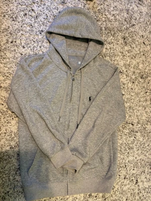 Grått loungewear-set från Polo Ralph Lauren - Mysigt loungewear-set i ljusgrått från Polo Ralph Lauren. Setet består av en hoodie med dragkedja och fickor samt matchande mjukisbyxor med resår i midjan och muddar vid bensluten. Båda plaggen har den klassiska Polo-loggan broderad på bröstet och benet.