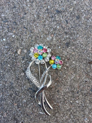 Blommig brosch med färgglada stenar - Snygg brosch i form av två blommor med glittrande stenar i flera färger som rosa, blå, grön, gul och vit. Metallbasen är silverfärgad och dekorerad med små klara stenar på bladen. Perfekt accessoar för att piffa upp din outfit.