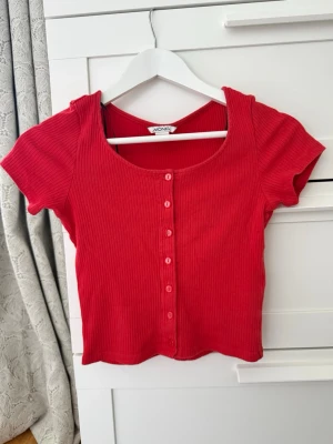 Röd ribbad topp från Monki - Kortärmad röd topp från Monki med ribbad struktur och knappar framtill. Toppen har en rundad urringning och croppad passform som ger en trendig look. Perfekt att matcha med höga jeans eller kjol.