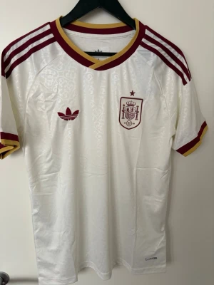 Vit Spanien fotbollströja finns i både storlek S och M  - En helt ny snygg vit fotbollströja från Adidas med Spaniens landslagsemblem på bröstet. Tröjan har vinröda och gula detaljer, klassiska Adidas-ränder på axlarna och retroinspirerat mönster i tyget. Perfekt för dig som älskar fotboll och vill sticka ut på planen. Finns i både storlek S och M