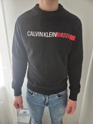 Svart sweatshirt Calvin Klein Jeans - Svart sweatshirt från Calvin Klein Jeans med vit och röd logga tryckt över bröstet. Tröjan har rund halsringning, ribbade muddar vid ärmslut och nederkant samt en klassisk, avslappnad passform. Perfekt till jeans för en clean och stilren look.