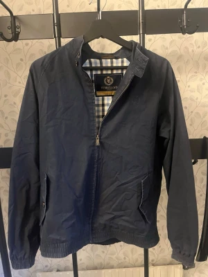 Marinblå overshirt från Henri Lloyd - Klassisk marinblå overshirt från Henri Lloyd med dragkedja och två sidofickor. Insidan har ett rutigt foder i blått och vitt. Overshirten har en enkel krage och elastiska muddar vid ärmslut och nederkant. Perfekt för en stilren och avslappnad look.