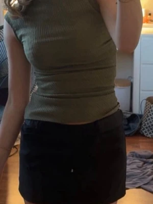 Olivgrön ribbad ärmlös topp - Snygg olivgrön ribbad topp med hög hals och ärmlös design. Perfekt att matcha med kjol eller jeans för en trendig look. Materialet är stretchigt och följsamt för en bekväm passform.