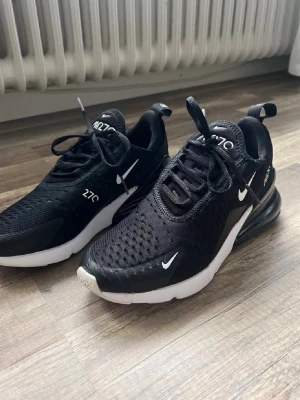Nike Air Max 270 svarta sneakers - Nike Air Max 270 sneakers i svart mesh och syntetmaterial med vita detaljer och synlig Air-enhet i hälen. Skorna har en sportig design, vita loggor och snörning. Perfekta för dig som gillar streetwear och vill ha bekväma, snygga sneakers.