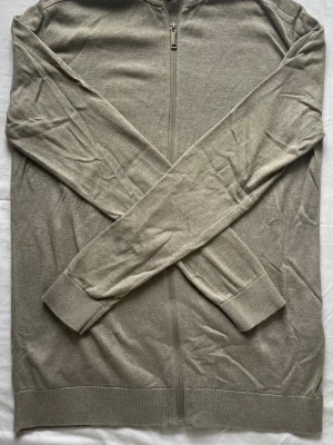 Zip tröja  - Stilren beige ziptröja med lång ärm och ribbade muddar. Tröjan har hel dragkedja framtill och är tillverkad i mjukt, tunt bomullsmaterial. Perfekt för lager på lager och enkel att matcha med olika outfits.