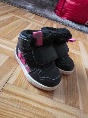 Svarta barnkängor med rosa detaljer - Säljer ett par svarta kängor för barn med rosa detaljer och fluffigt foder. Kardborreband gör dem enkla att ta på och av. Yttermaterialet är i syntet och textil, sulan är vit med rosa inslag. Perfekta för kalla dagar.