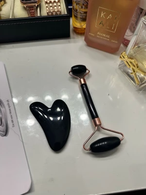 Svart ansiktsroller och gua sha-set - Snyggt set med svart ansiktsroller och gua sha-sten. Rollern har metalliska detaljer i roséguld och två släta rullar i svart sten, perfekt för ansiktsmassage. Gua sha-stenen är hjärtformad och tillverkad i samma svarta material för skönhetsrutinen.
