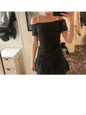 Svart offshoulder kortklänning - Snygg svart offshoulder klänning med kort ärm och volang nedtill. Klänningen har en figurnära passform och är perfekt för dig som gillar en stilren look. Materialet känns mjukt och följsamt. Strlk L men passar mindre