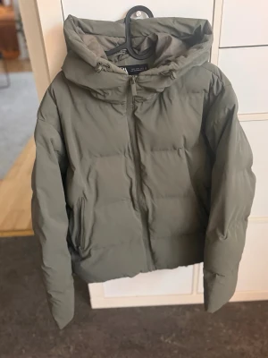 Olivgrön pufferjacka med huva - Snygg olivgrön pufferjacka med dragkedja framtill och två sidofickor. Jackan har en stor huva och quiltad design som ger en modern look. Perfekt för kalla dagar och enkel att matcha med olika outfits. Materialet känns mjukt och isolerande.